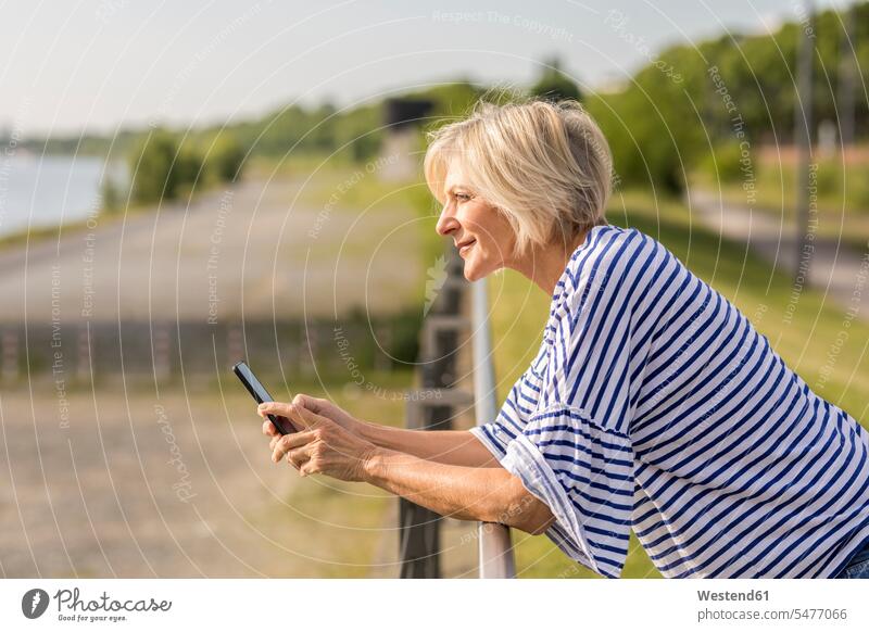 Lächelnde ältere Frau mit Handy, die sich am Flussufer an ein Geländer lehnt Mobiltelefon Handies Handys Mobiltelefone weiblich Frauen lächeln aufstützen