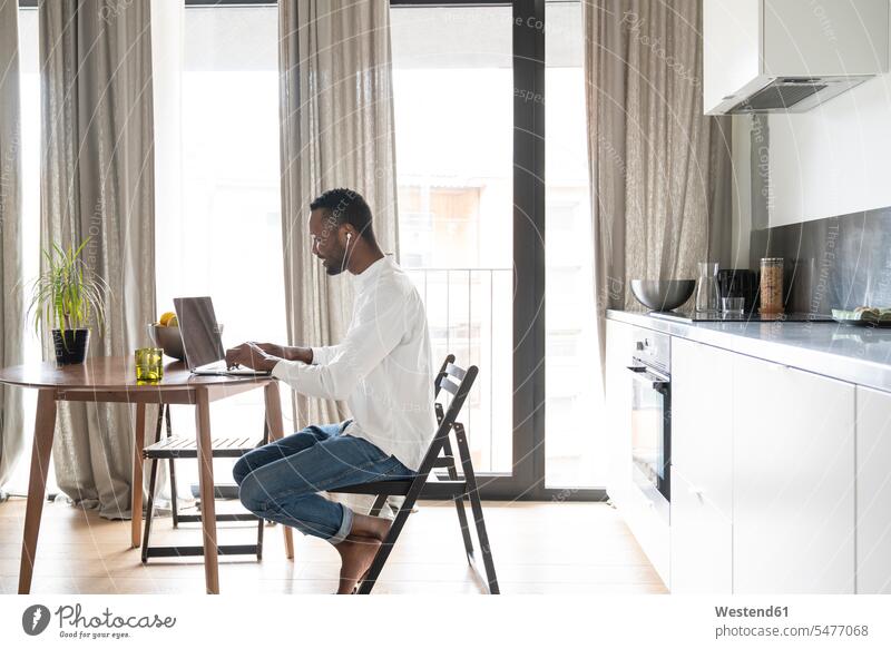 Lächelnder Mann sitzt in moderner Wohnung mit Laptop und Kopfhörern am Tisch Tische Holztische Rechner Laptops Notebook Notebooks sitzend daheim zu Hause
