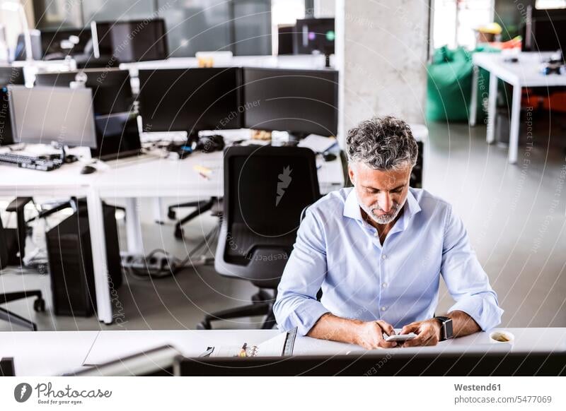 Reifer Geschäftsmann sitzt am Schreibtisch im Büro und benutzt ein Smartphone Arbeitstisch Schreibtische sitzen sitzend iPhone Smartphones Office Büros