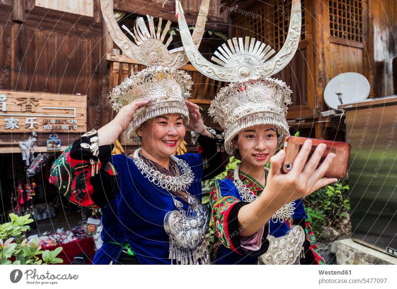 China, Guizhou, zwei Miao-Frauen in traditionellen Kleidern und mit Kopfschmuck machen ein Selfie mit Smartphone Bergdorf Ritual Rituale iPhone Smartphones