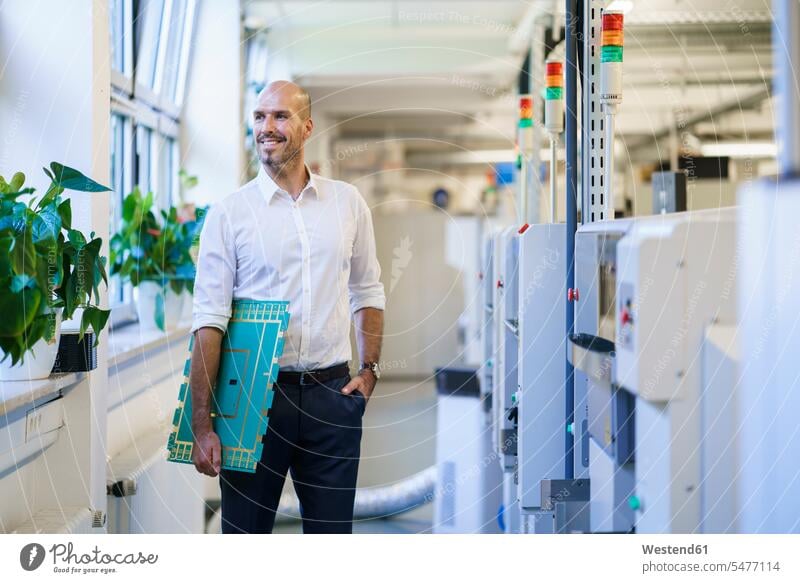 Lächelnder männlicher Ingenieur schaut weg, während er eine große Leiterplatte an Maschinen in der Fabrik hält Farbaufnahme Farbe Farbfoto Farbphoto