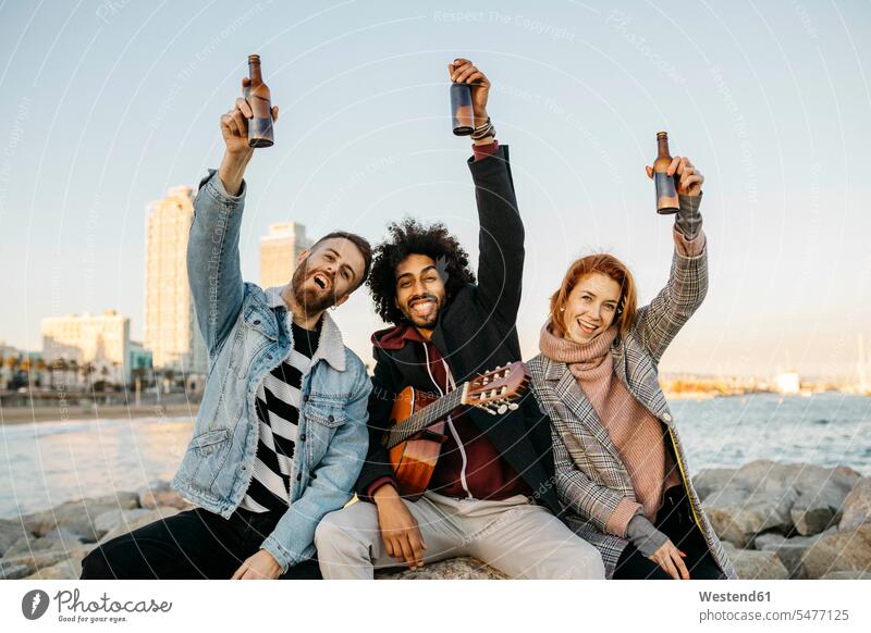 Porträt von drei glücklichen Freunden mit Gitarre heben Bierflaschen an der Küste Sonnenuntergang erheben Kueste Kuesten Küsten Glück glücklich sein