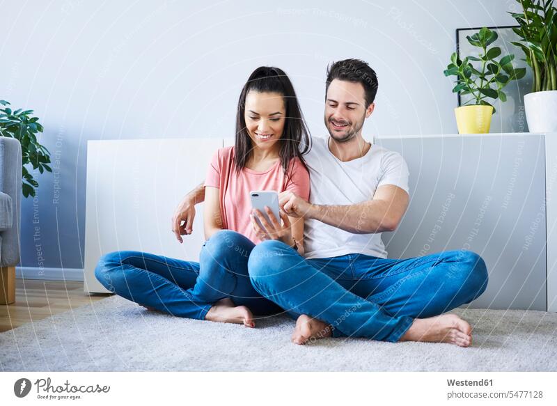 Paar, das auf dem Boden sitzt und ein Smartphone benutzt iPhone Smartphones sitzen sitzend Pärchen Paare Partnerschaft Böden Boeden Handy Mobiltelefon Handies