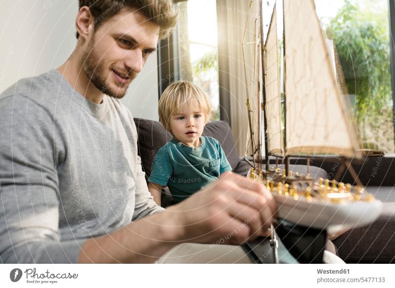 Vater und Sohn schauen sich zu Hause auf der Couch ein Spielzeug-Modellschiff an sitzen sitzend sitzt ansehen Zuhause daheim Modellboot Sofa Couches Liege Sofas