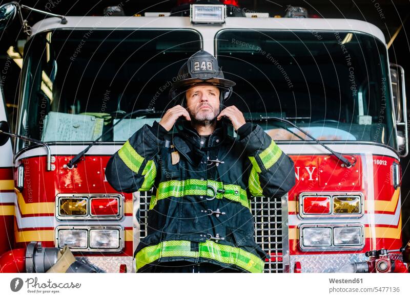USA, New York, Porträt eines Feuerwehrmannes, der einen Helm vor einem Feuerwehrauto aufsetzt Vereinigte Staaten von Amerika United States of America Portrait