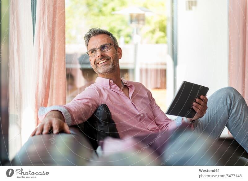 Lächelnder reifer Mann sitzt zu Hause auf der Couch und hält eine Tablette sitzen sitzend lächeln Sofa Couches Liege Sofas Männer männlich Tablet Computer