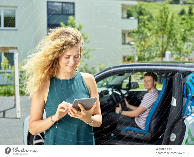 Lächelnde junge Frau benutzt Tablette mit einem Mann, der im Hintergrund im Elektroauto sitzt Auto Wagen PKWs Automobil Autos lächeln Paar Pärchen Paare