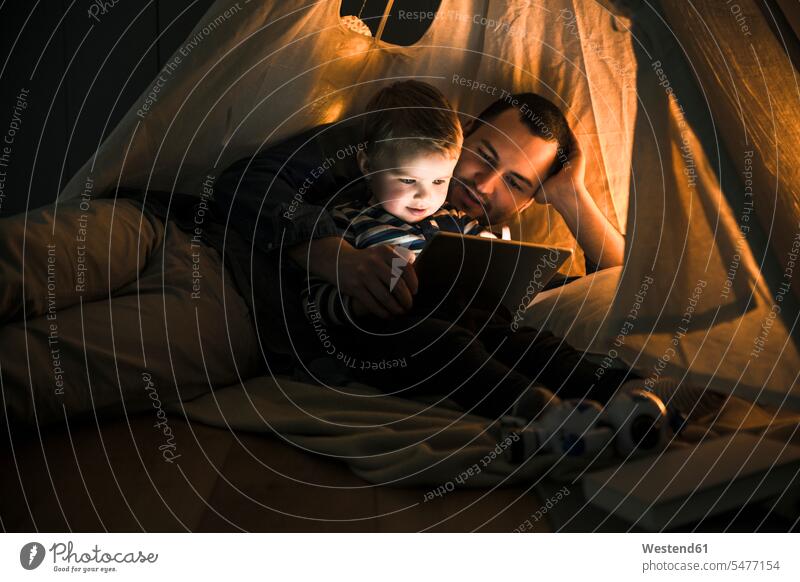 Vater und Sohn teilen sich ein Tablet in einem dunklen Zelt zu Hause dunkel Zuhause daheim Zelte Teilen Sharing liegen liegend liegt Papas Väter Vati Vatis