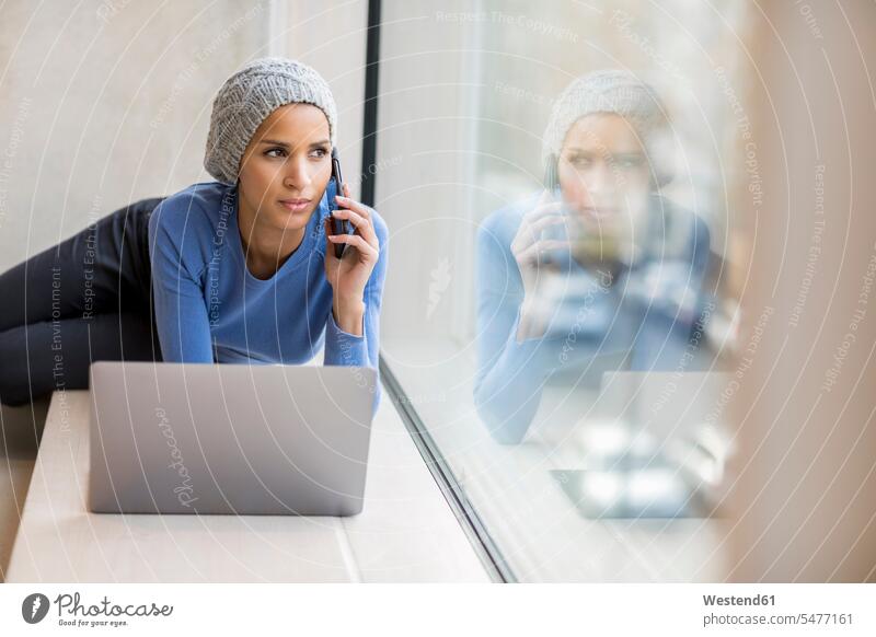 Porträt einer jungen Frau am Telefon mit Laptop auf dem Fensterbrett, die aus dem Fenster schaut schauen schauend anschauen betrachten Notebook Laptops