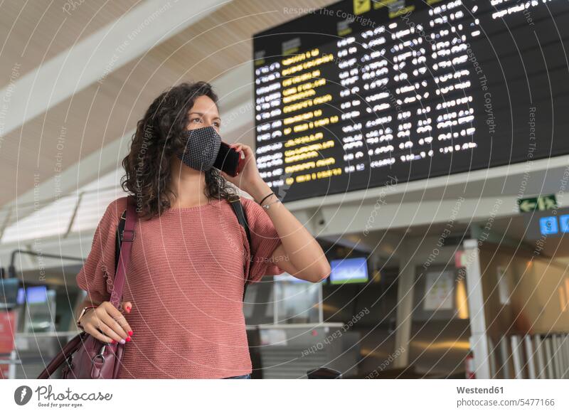 Junge Frau mit schützender Gesichtsmaske telefoniert im Stehen am Flughafen mit einem Smartphone Farbaufnahme Farbe Farbfoto Farbphoto Innenaufnahme