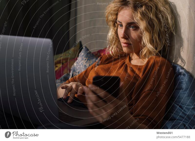 Porträt einer blonden jungen Frau, die mit Handy und Laptop auf dem Bett liegt Leute Menschen People Person Personen Europäisch Kaukasier kaukasisch 1 Ein
