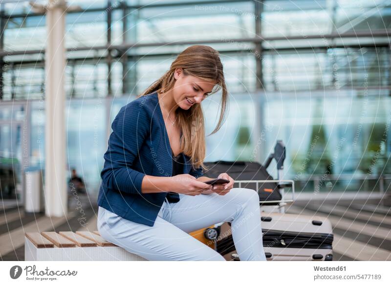 Lächelnde junge Geschäftsfrau sitzt im Freien mit Handy und Koffer Mobiltelefon Handies Handys Mobiltelefone Geschäftsfrauen Businesswomen Businessfrauen