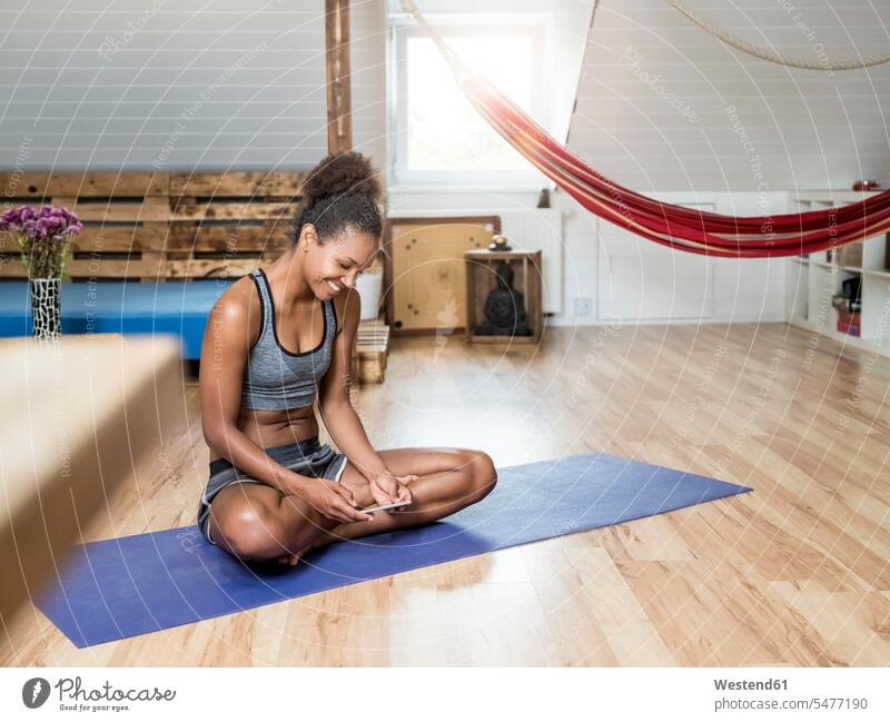 Lächelnde junge Frau sitzt auf Yogamatte und schaut auf Handy weiblich Frauen Turnmatte Sportmatte Yoga-Matte ansehen Mobiltelefon Handies Handys Mobiltelefone