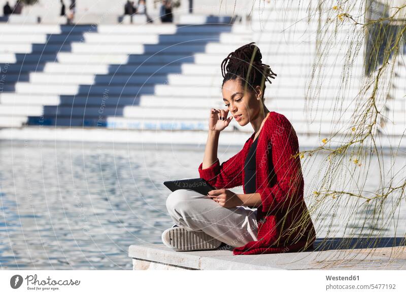 Junge Frau sitzt im Schneidersitz und benutzt ein digitales Tablet, während sie am Rande der Promenade über den Fluss an einem sonnigen Tag steht Farbaufnahme
