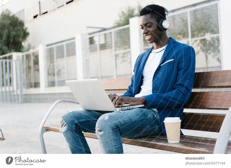 Lächelnder junger Geschäftsmann, der einen Laptop benutzt, während er auf einer Bank am Fußweg Musik hört Farbaufnahme Farbe Farbfoto Farbphoto Außenaufnahme