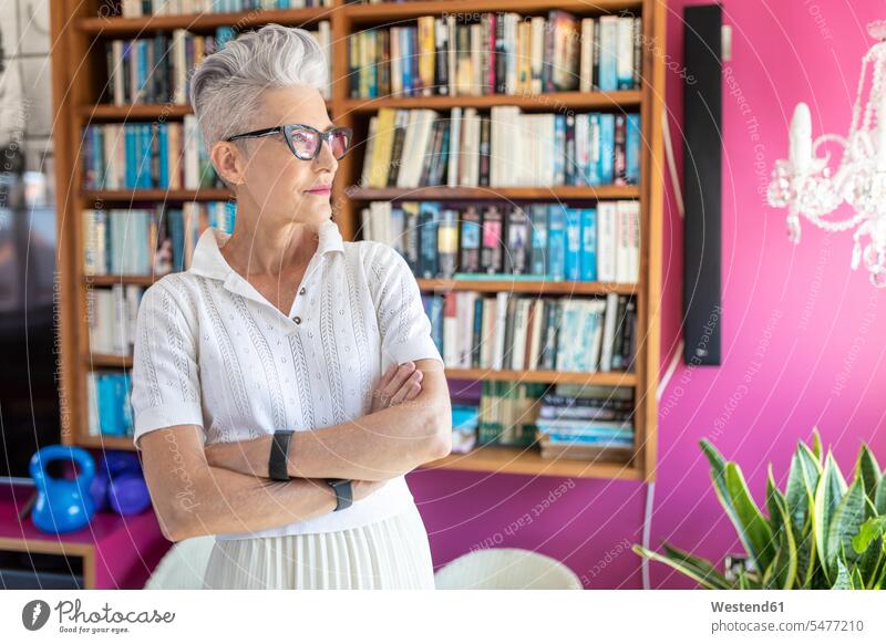 Ältere Frau mit verschränkten Armen, die zu Hause gegen ein Bücherregal steht Farbaufnahme Farbe Farbfoto Farbphoto Tag Tageslichtaufnahme Tageslichtaufnahmen