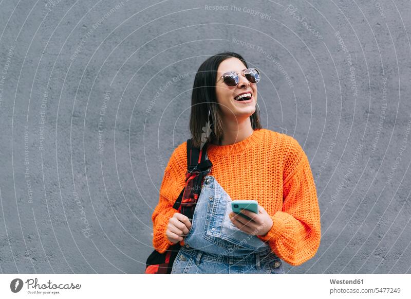 Lächelnde Frau mit Sonnenbrille und Geldbörse, die ein Mobiltelefon benutzt, während sie an einer grauen Wand steht Farbaufnahme Farbe Farbfoto Farbphoto