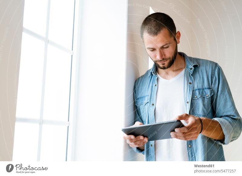 Junger ernster Mann zu Hause, der auf sein Tablet schaut, das am Fenster steht Leute Menschen People Person Personen kurzes Haar kurzes Haare Kurzhaarfrisur
