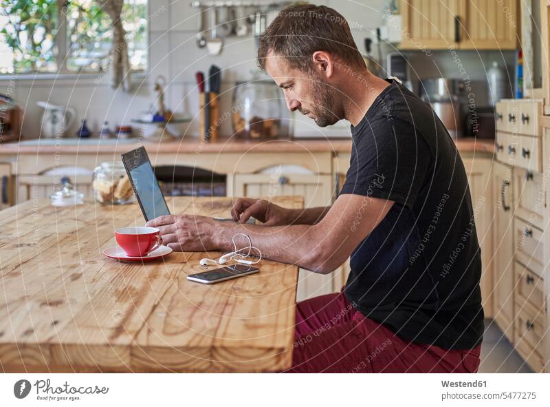 Mann arbeitet von zu Hause aus, sitzt am Küchentisch, benutzt Laptop und Smartphone Job Berufe Berufstätigkeit Beschäftigung Jobs Tische Holztische Rechner