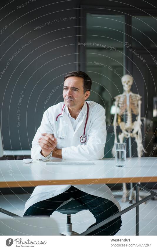 Arzt sitzt am Schreibtisch in der Arztpraxis mit Skelett im Hintergrund Skelette Doktoren Ärzte Arztpraxen Praxis Arbeitstisch Schreibtische sitzen sitzend