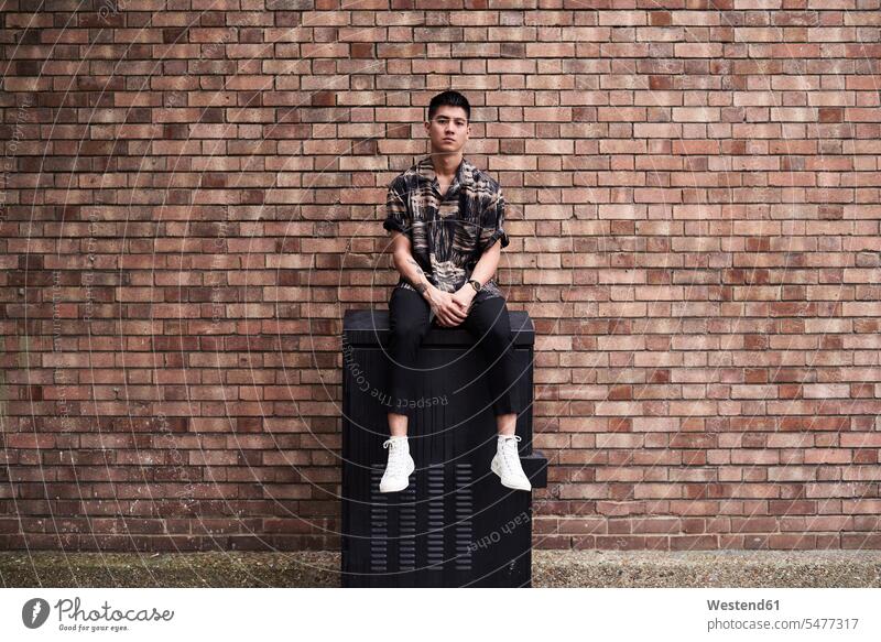 Porträt eines jungen Mannes vor einer Ziegelmauer Hemden sitzend sitzt Individuell Stil stylisch Koerperschmuck Taetowierung Taetowierungen Tatoo Tatoos Tattoos