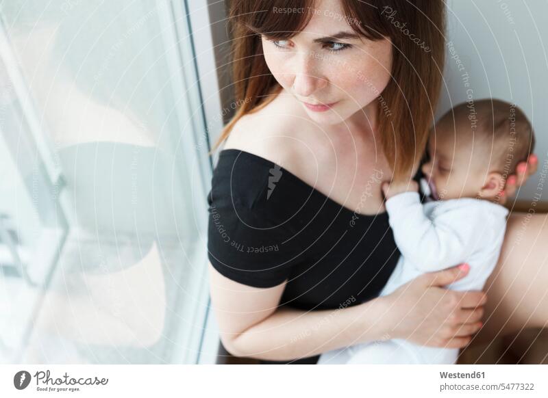 Junge Mutter mit ihrem kleinen Mädchen, die zu Hause aus dem Fenster schaut sitzend sitzt behüten behütet geborgen Sicherheit innig nah Erwartung Sehnsucht