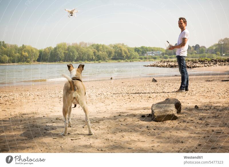 Mann mit Hund fliegt Drohne an einem Fluss Fluesse Fluß Flüsse Drohnen Männer männlich fliegen fliegend Hunde Gewässer Wasser Erwachsener erwachsen Mensch