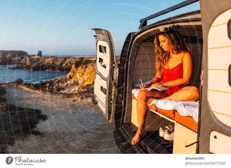 Frau schreibt in Buch, während sie im Wohnmobil gegen am Strand sitzt Farbaufnahme Farbe Farbfoto Farbphoto Außenaufnahme außen draußen im Freien