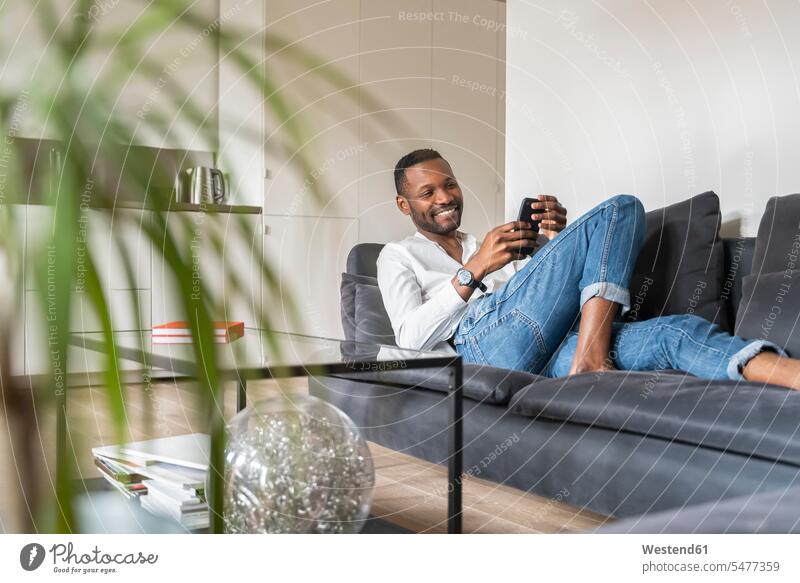 Porträt eines lächelnden Mannes, der in einer modernen Wohnung auf der Couch sitzt und ein Smartphone benutzt Uhren Armbanduhren Couches Liege Sofas