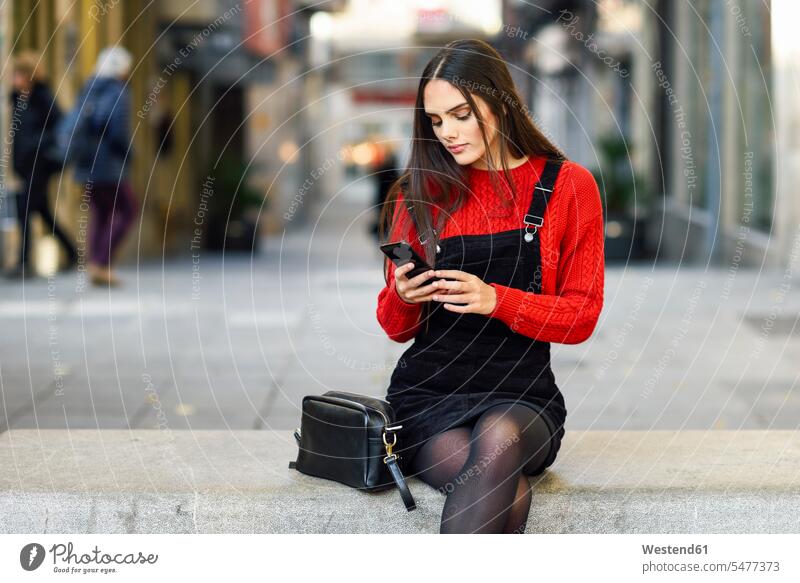 Porträt einer modischen jungen Frau, die auf einer Bank in der Fußgängerzone sitzt und auf ihr Handy schaut sitzen sitzend weiblich Frauen Smartphone iPhone