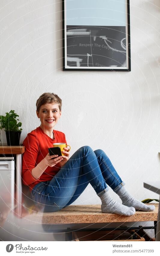 Frau sitzt in der Küche, trinkt Kaffee und hört Smartphone-Nachrichten ab trinken entspannt entspanntheit relaxt weiblich Frauen glücklich Glück glücklich sein