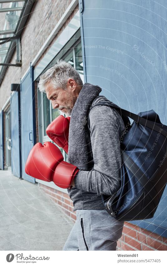 Älterer Mann mit Handtuch, Sporttasche und roten Boxhandschuhen vor einem Fitnessstudio stehend Handtuecher Handtücher Männer männlich Fitnessclubs
