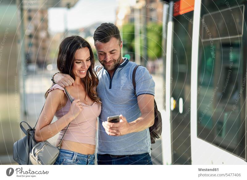 Ehepaar schaut auf ein Smartphone, während es am Bahnhof auf die Straßenbahn wartet Handy Mobiltelefon Handies Handys Mobiltelefone Station Haltestelle