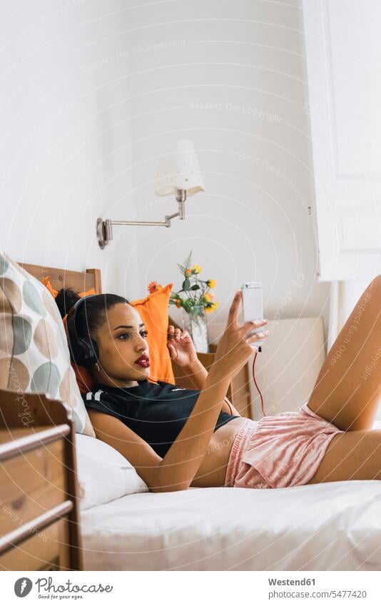 Schöne junge Frau liegt zu Hause auf dem Bett, trägt Kopfhörer und schaut auf ihr Handy Zuhause daheim ansehen Betten Kopfhoerer Mobiltelefon Handies Handys