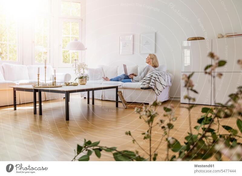 Blonde, reife Frau sitzt auf der Couch im Wohnzimmer und benutzt einen Laptop Sofa Couches Liege Sofas sitzen sitzend blond blonde Haare blondes Haar weiblich