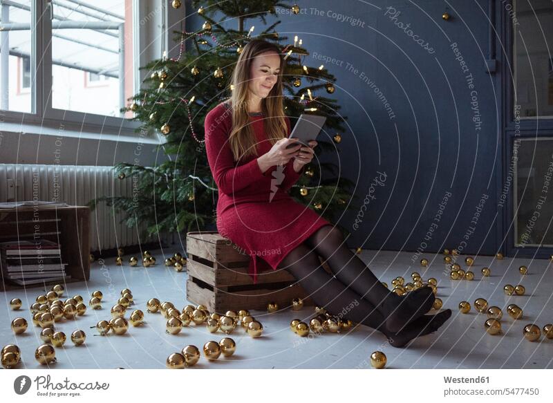 Lächelnde Frau sitzt auf Holzkiste vor geschmücktem Weihnachtsbaum mit Tablette weiblich Frauen sitzen sitzend Tablet Computer Tablet-PC Tablet PC iPad