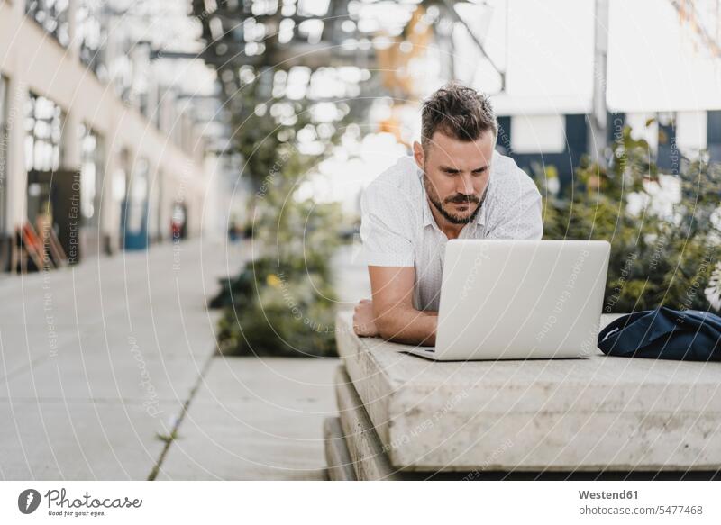 Junger Mann mit Laptop und Blick zur Seite, auf einer Bank in der Stadt liegend Job Berufe Berufstätigkeit Beschäftigung Jobs geschäftlich Geschäftsleben