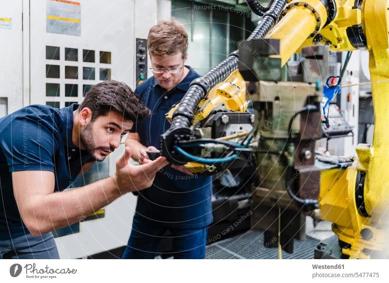 Männliche Mitarbeiter untersuchen Roboterarm, während sie in der Fabrik stehen Farbaufnahme Farbe Farbfoto Farbphoto Innenaufnahme Innenaufnahmen innen drinnen