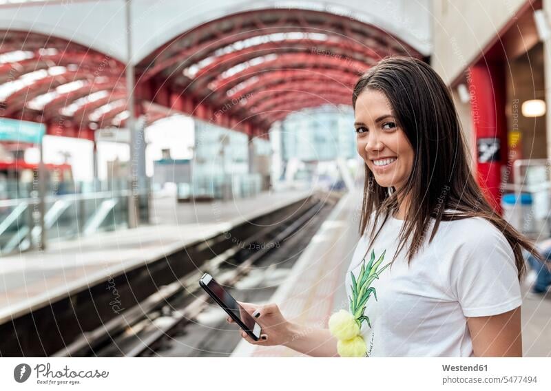UK, London, Porträt einer lächelnden jungen Frau, die ein Handy in einer Bahnstation hält Portrait Porträts Portraits Mobiltelefon Handies Handys Mobiltelefone