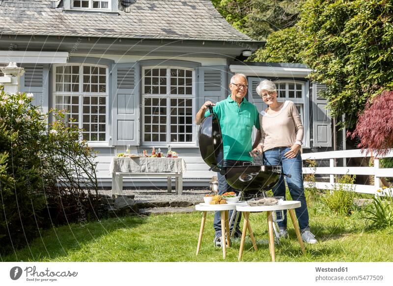 Glückliches älteres Ehepaar beim Grillen im Garten ihres Hauses Tische Grillgeraet Grillgeraete Grillgerät Grillgeräte Grills Brillen sommerlich Sommerzeit