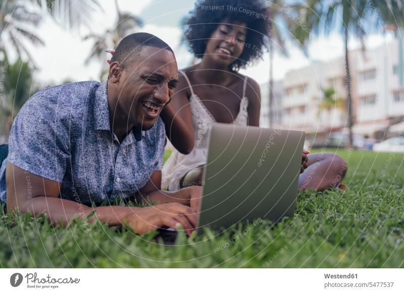 USA, Florida, Miami Beach, lachendes junges Paar schaut auf Laptop auf Rasen in einem Park ansehen Notebook Laptops Notebooks Parkanlagen Parks Pärchen Paare