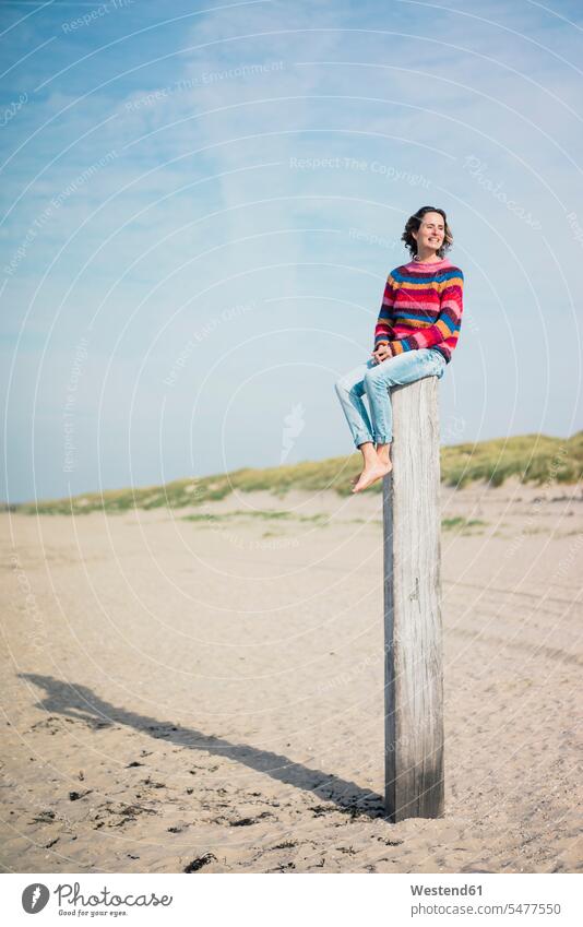 Reife Frau sitzt auf einem Holzpfahl am Strand sitzen sitzend Unabhängigkeit selbständig Selbständigkeit Beach Straende Strände Beaches Holzpfosten Niederlande