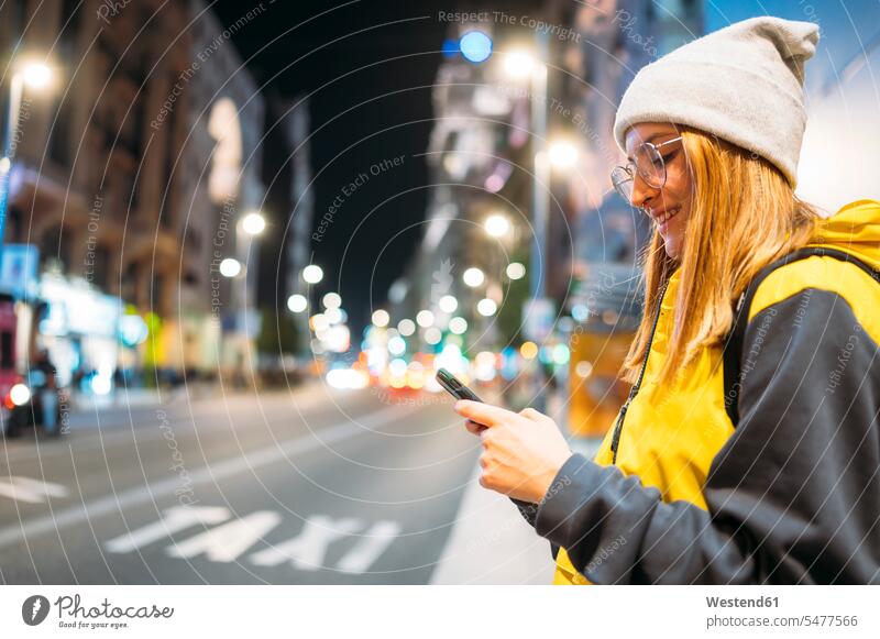 Lächelnde junge Frau in der Stadt, die nachts ihr Smartphone benutzt Rucksäcke KFZ Verkehrsmittel Automobil Autos PKW PKWs Wagen Telekommunikation telefonieren