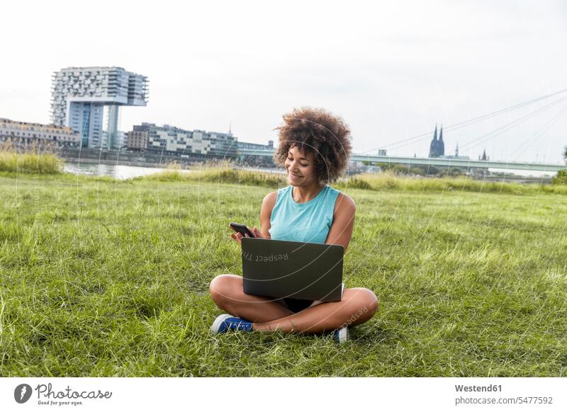 Deutschland, Köln, Frau sitzt auf der Wiese und benutzt Laptop und Mobiltelefon weiblich Frauen Notebook Laptops Notebooks Handy Handies Handys Mobiltelefone