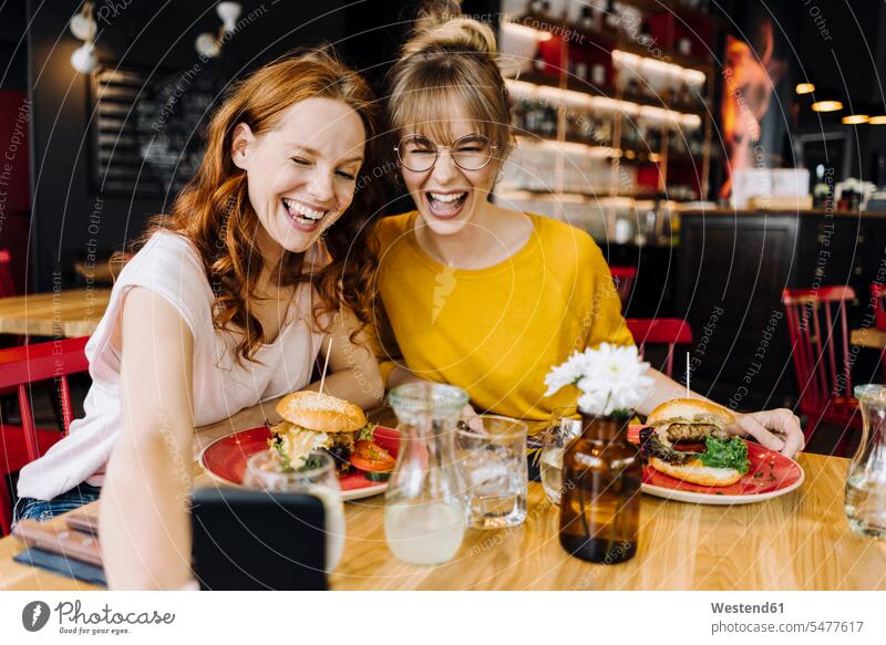 Zwei glückliche Freundinnen essen einen Burger und machen ein Selfie in einem Restaurant Telekommunikation telefonieren Handies Handys Mobiltelefon