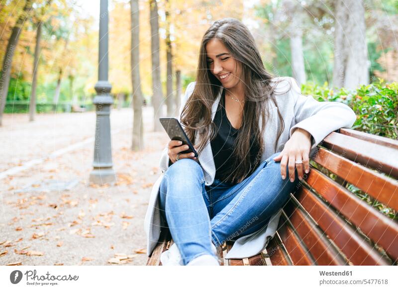 Lächelnde Frau sitzt auf einer Parkbank und benutzt ein Smartphone Bänke Sitzbank Sitzbänke Parkbänke Telekommunikation telefonieren Handies Handys Mobiltelefon
