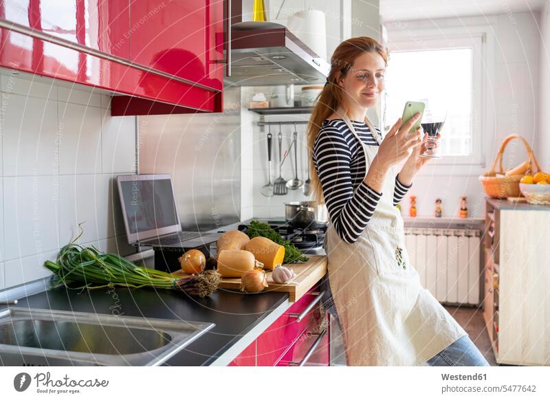 Frau trinkt ein Glas Rotwein in der Küche, während sie ein Smartphone benutzt Leute Menschen People Person Personen erwachsen jung Millennial 20 - 30 Jahre