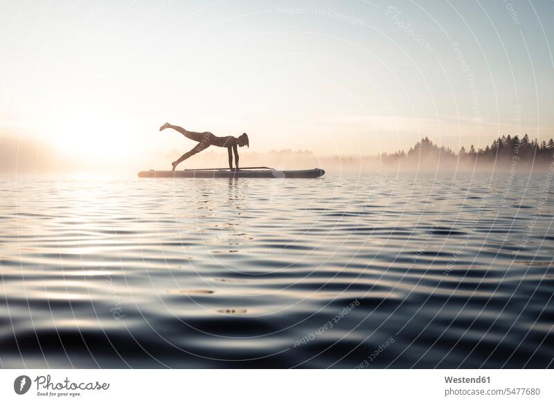 Frau praktiziert morgens Paddelbrett-Yoga auf dem Kirchsee, Bad Tölz, Bayern, Deutschland ausüben trainieren Übung früh Frühe Morgen geniessen Genuss