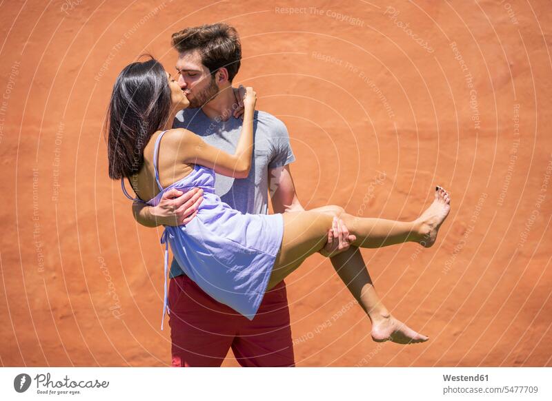 Junger Mann küsst, während er eine Frau bei Sonnenschein gegen eine braune Wand im Hinterhof trägt Farbaufnahme Farbe Farbfoto Farbphoto Spanien Zusammenhalt