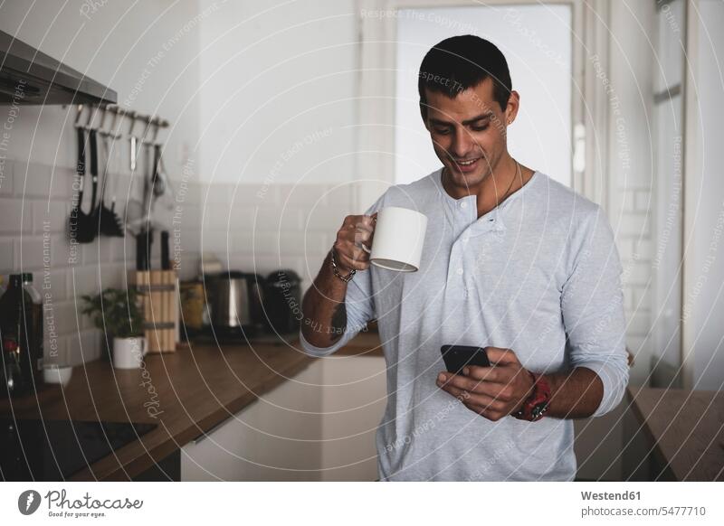 Lächelnder junger Mann mit Tasse Kaffee mit Handy in der Küche zu Hause Männer männlich Zuhause daheim lächeln Kaffeetasse Kaffeetassen Mobiltelefon Handies